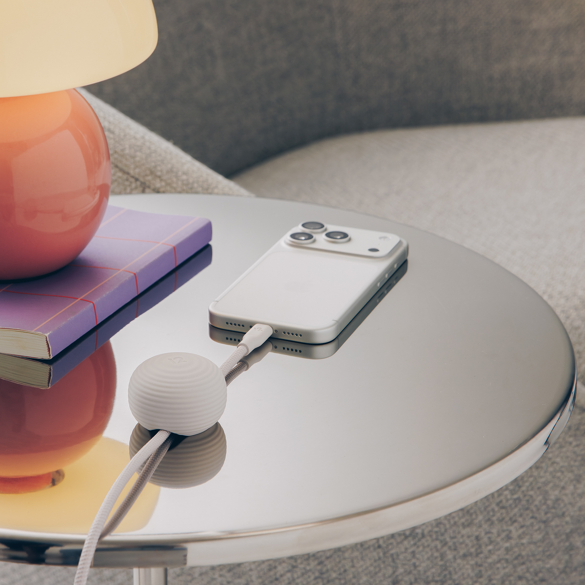 Smartphone with a charging cable on a glass table next to a lamp and books.