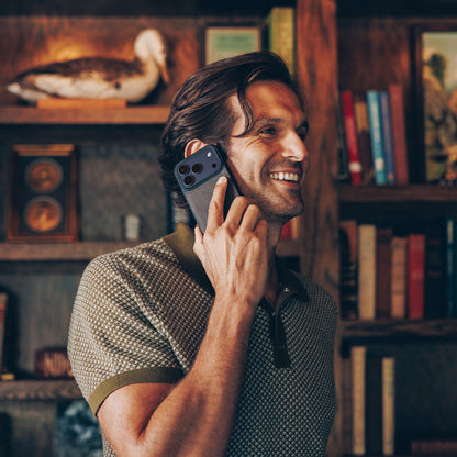 Man talking on a phone in a cozy room with bookshelves and decorative items.