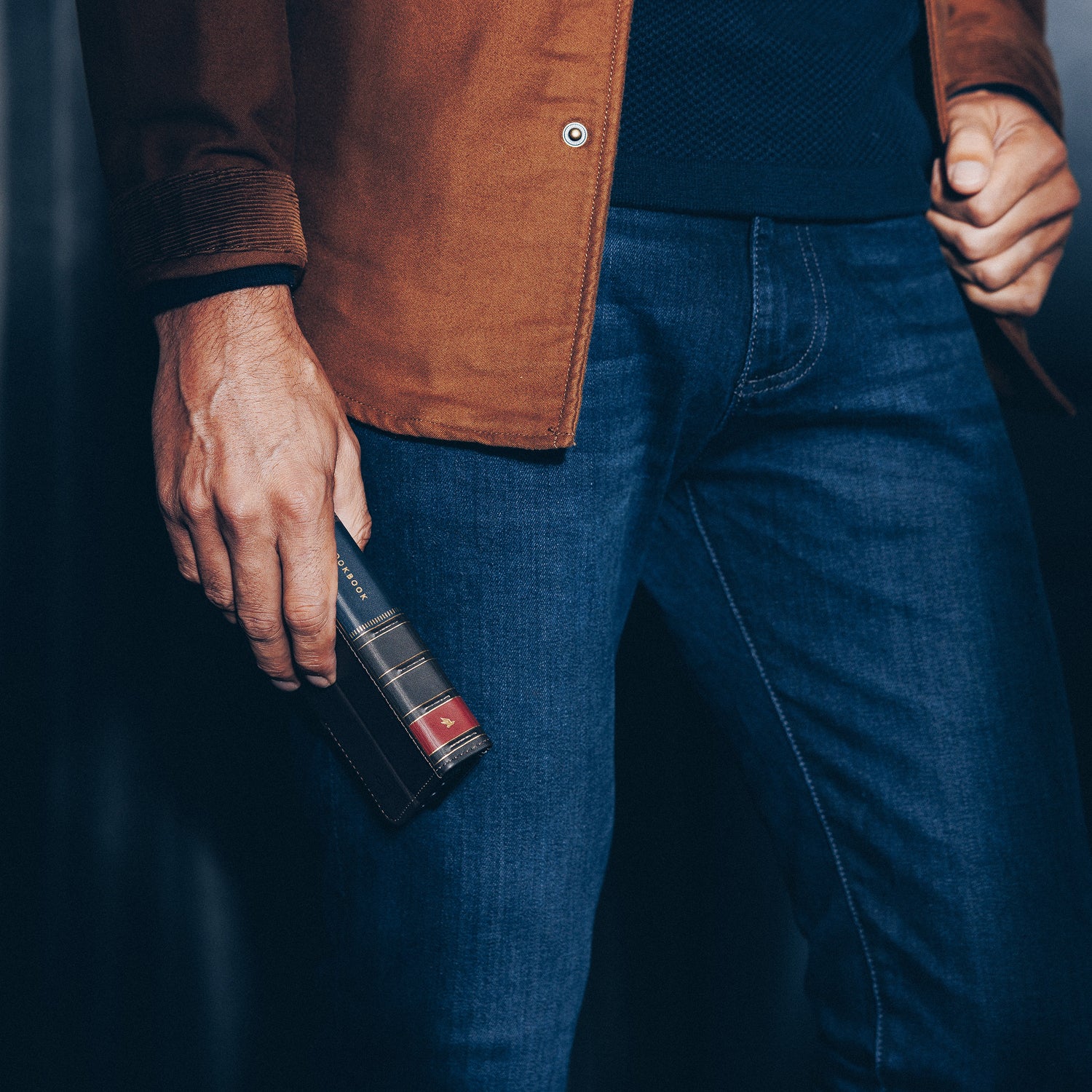 Person wearing a brown jacket and blue jeans holding a book.