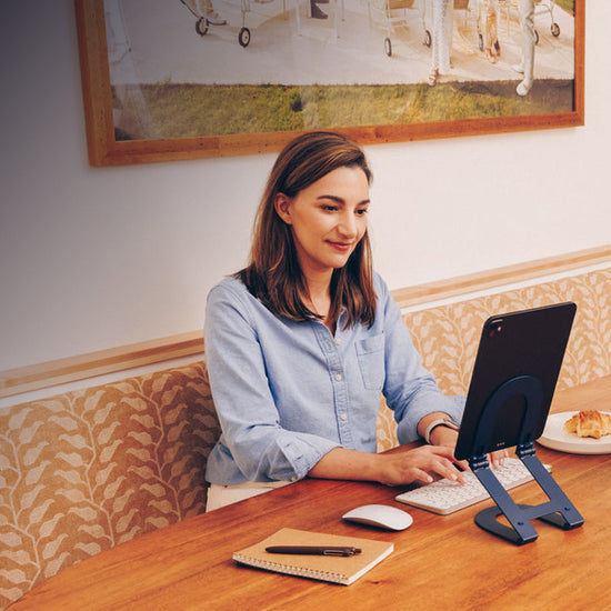 Woman using a tablet on a stand sitting at a wooden table with a notebook and pen in a cozy room.
