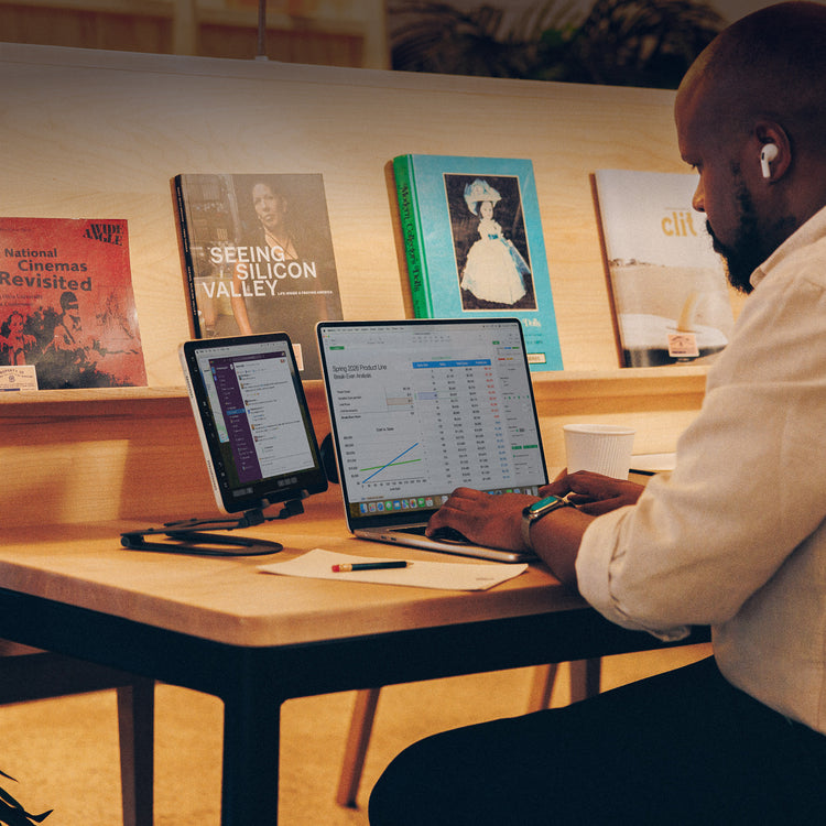 Person working on a laptop and tablet at a table with books and a cup in the background